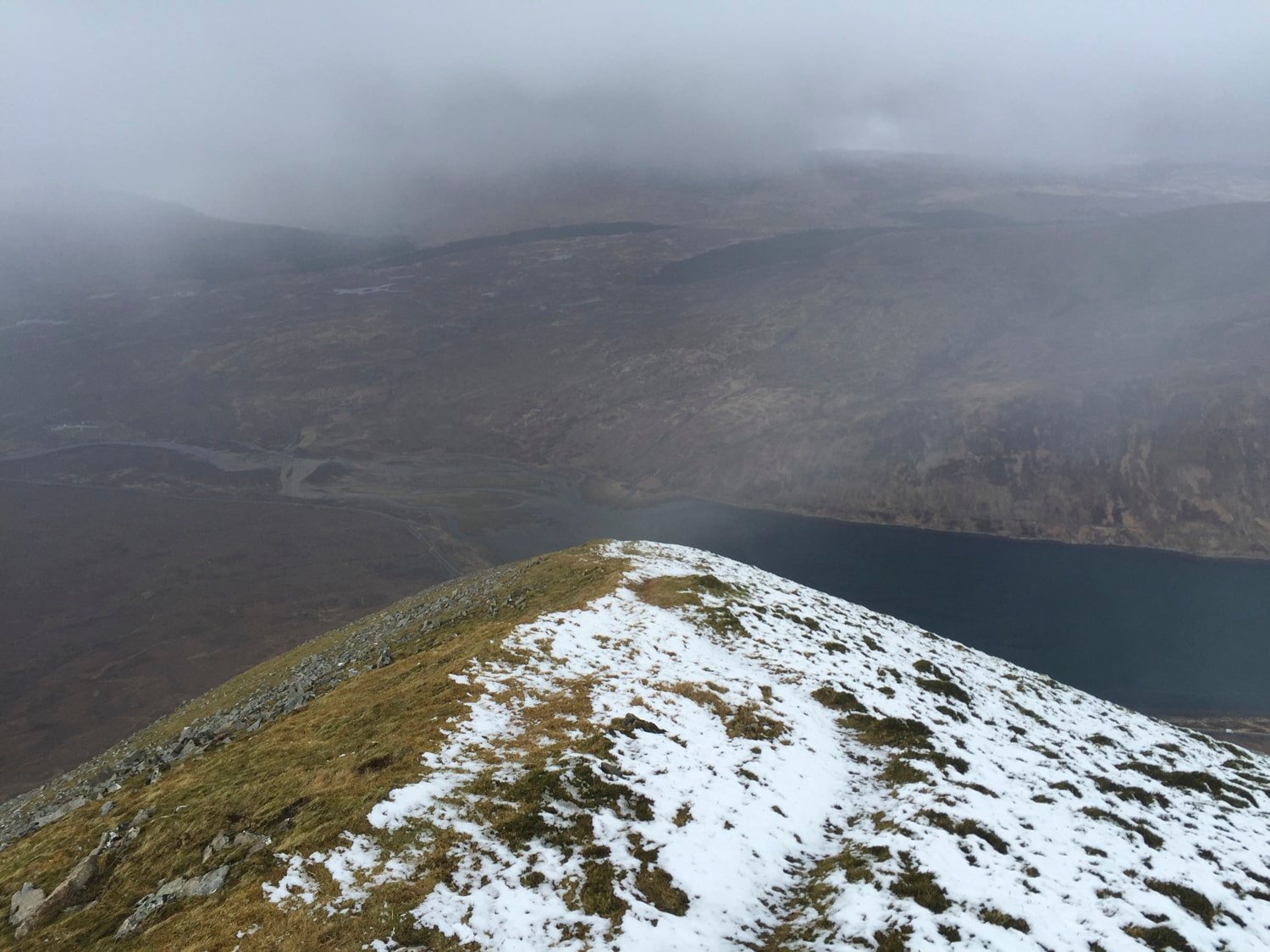 Vue depuis le sommet — crête enneigée, loch sombre en contrebas, brume